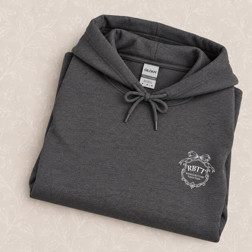 Raised Better Than This crest hoodie in Dark Heather folded on a cream floral surface, crest close up