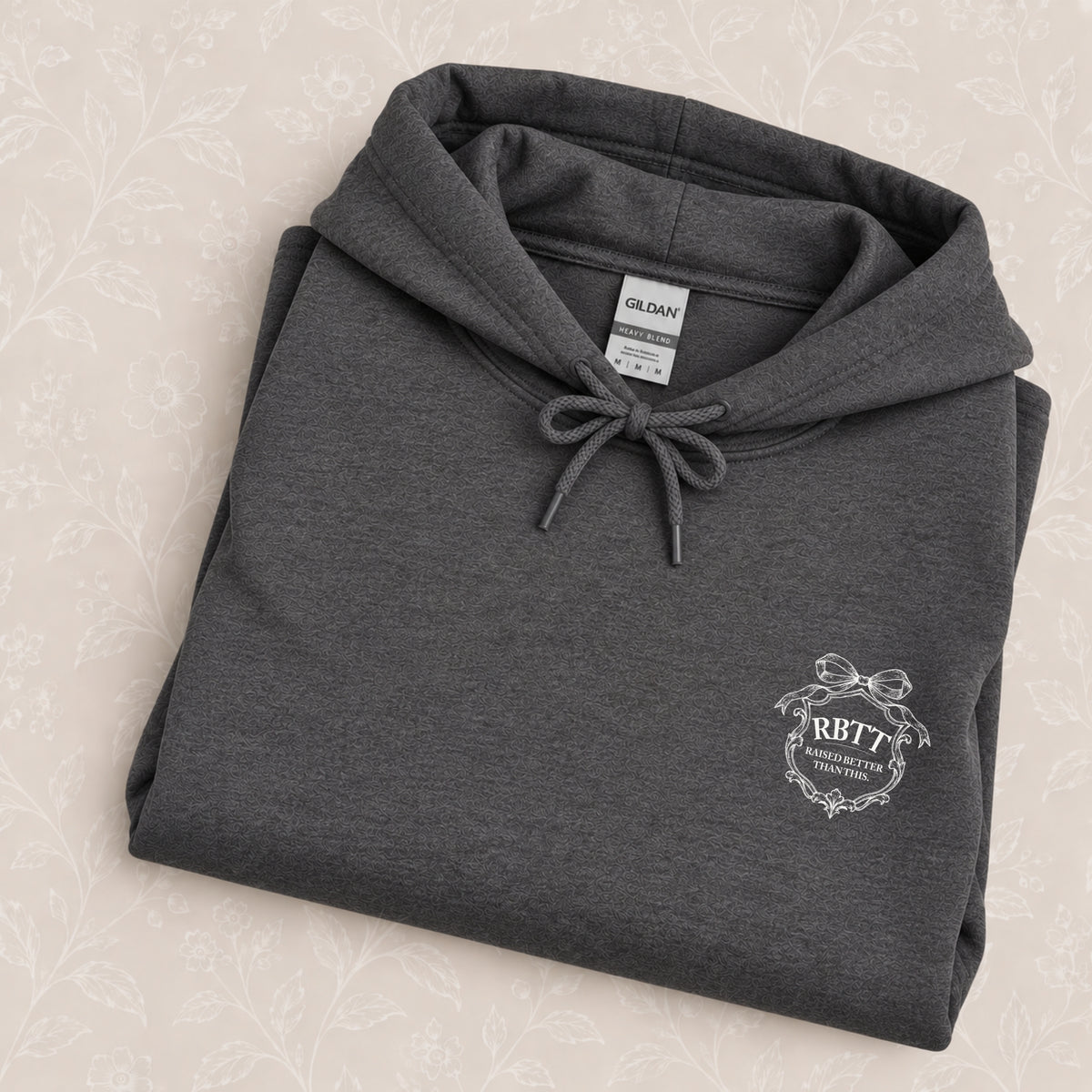 Raised Better Than This crest hoodie in Dark Heather folded on a cream floral surface, crest close up