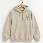 Raised Better Than This crest hoodie in Sand on a wooden hanger, navy left-chest crest