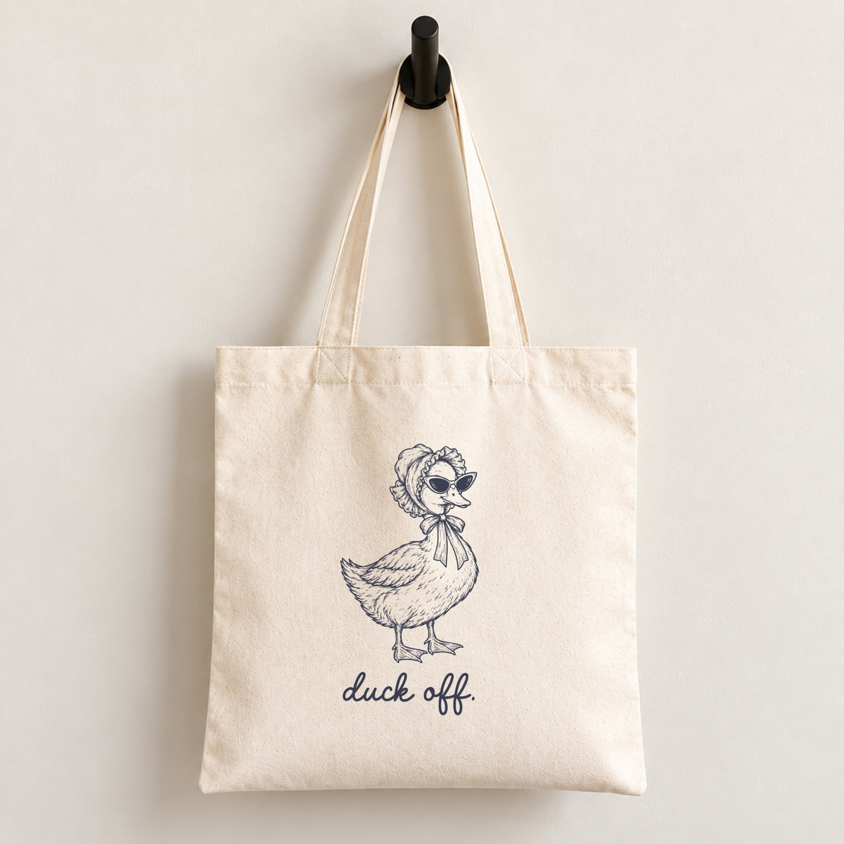Duck Off natural canvas tote bag hanging on a wall hook, navy duck in a bonnet and sunglasses printed on the front