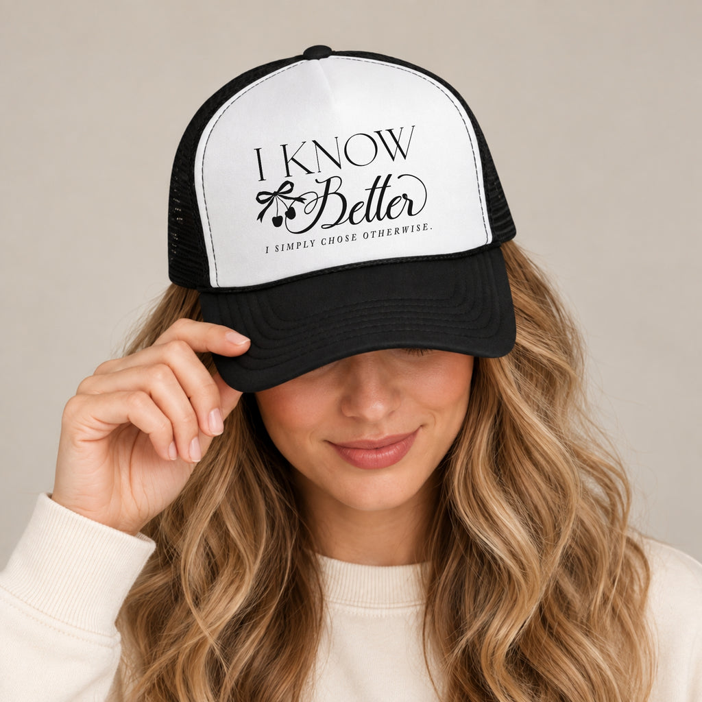 Woman in a cream sweatshirt tipping the brim of the I Know Better trucker hat in Black with a white front panel