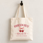 Poor Judgment Society Lifetime Member canvas tote bag hanging on a wall hook, cherry bow crest in deep red