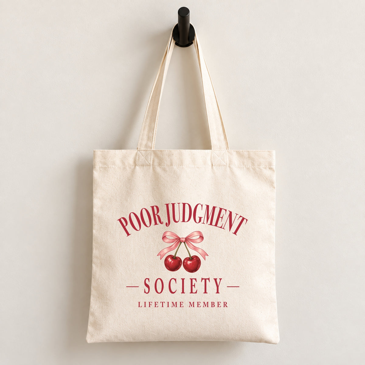 Poor Judgment Society Lifetime Member canvas tote bag hanging on a wall hook, cherry bow crest in deep red