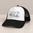 I Know Better trucker hat in Black on a table, angled view showing the mesh back and curved bill