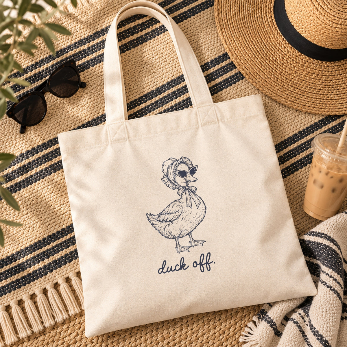 Duck Off canvas tote flat lay with a straw hat, sunglasses and an iced coffee on a striped blanket