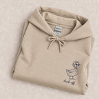 Duck Off hoodie in Sand folded on a cream floral surface, close up of the duck in a bonnet and cat-eye sunglasses