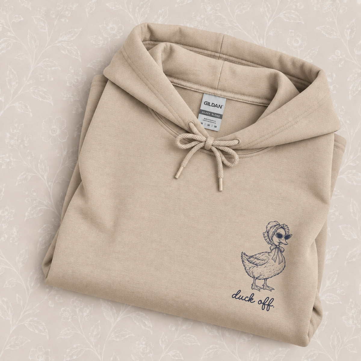Duck Off hoodie in Sand folded on a cream floral surface, close up of the duck in a bonnet and cat-eye sunglasses