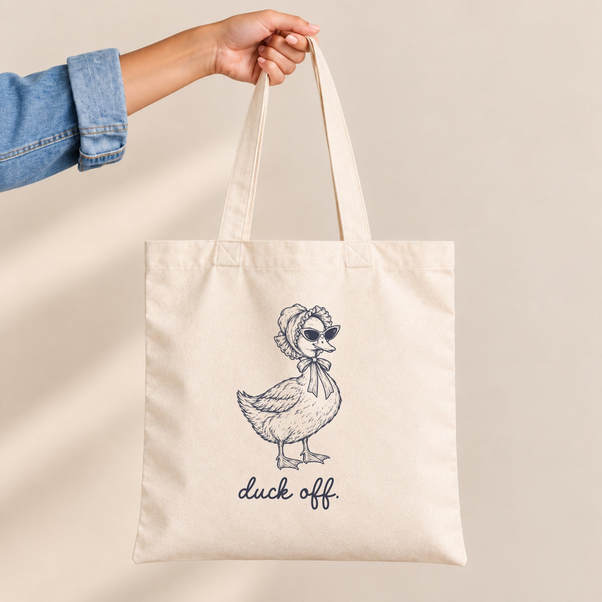 Hand holding the Duck Off natural canvas tote by its long handles, duck design close up