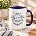 Raised Better Than This crest mug with navy handle on a marble counter with a knit throw, plant and candle