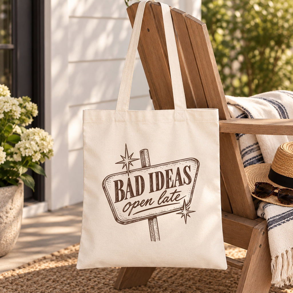 Bad Ideas, Open Late canvas tote bag hanging on a porch chair with a striped blanket and sun hat