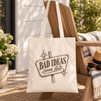 Bad Ideas, Open Late canvas tote bag hanging on a porch chair with a striped blanket and sun hat