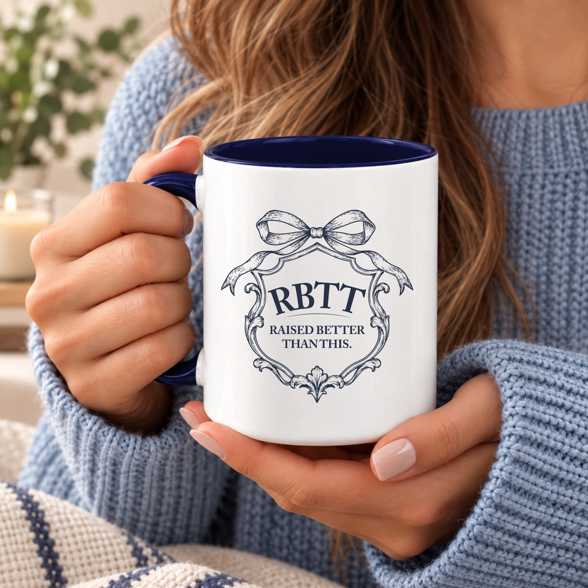 Woman in a blue knit sweater holding the Raised Better Than This crest mug in both hands