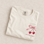 Mostly Together cherry bow tee in Ivory folded on a cream floral surface, left-chest print close up