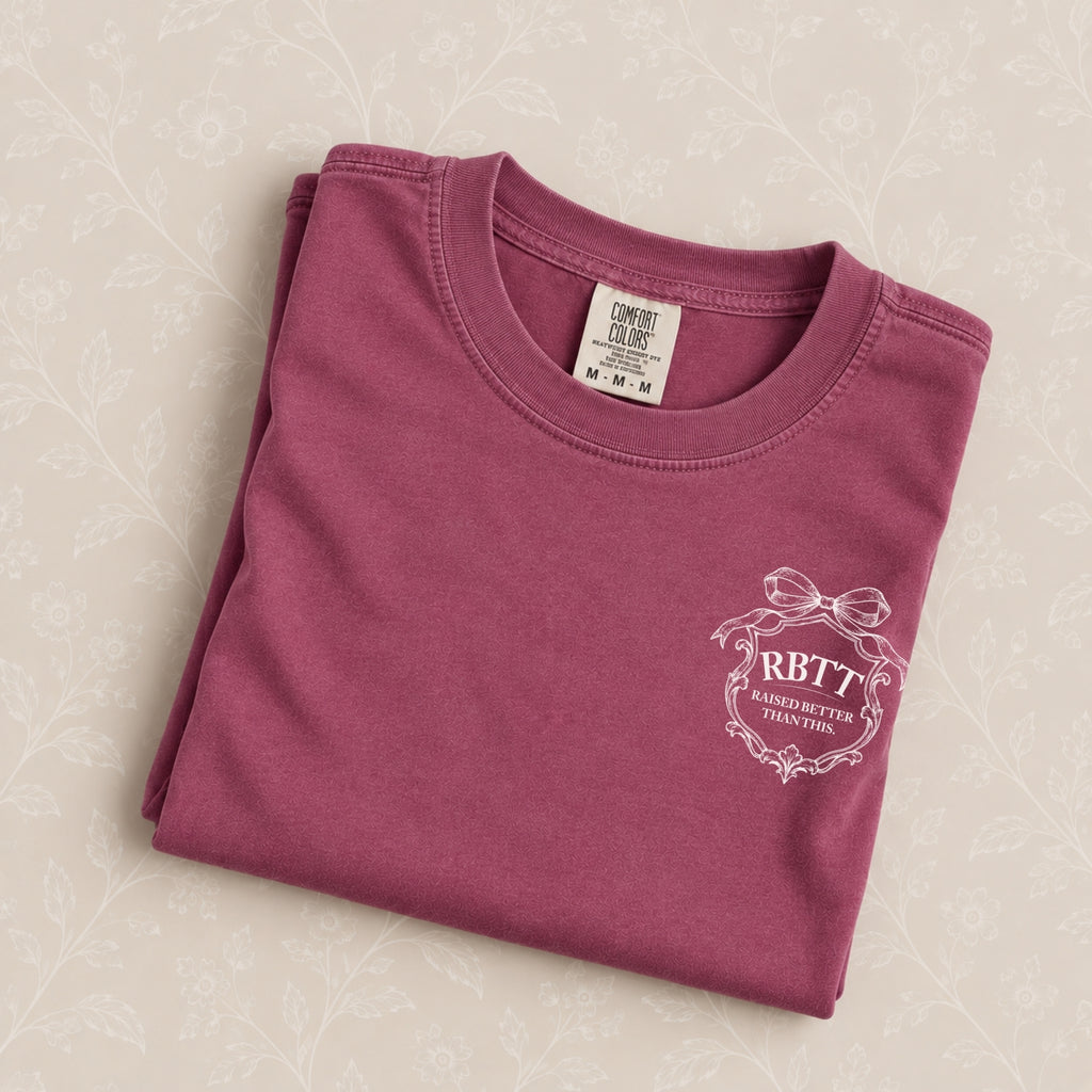 Raised Better Than This crest tee in Berry, folded on a cream floral surface, RBTT logo close up
