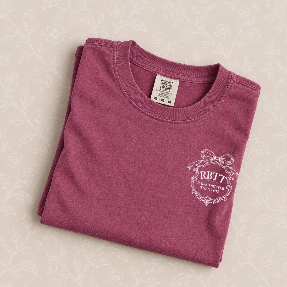 Raised Better Than This crest tee in Berry, folded on a cream floral surface, RBTT logo close up