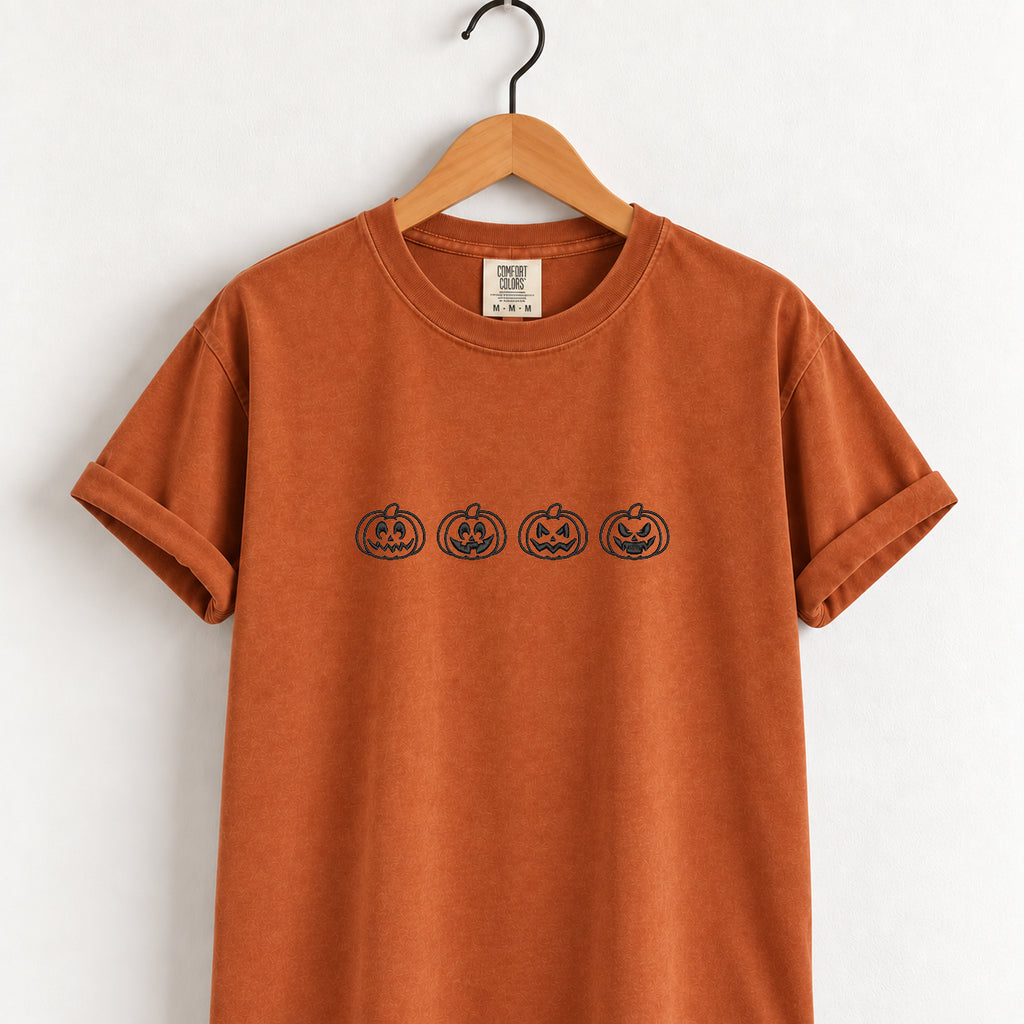 Jack-o-lantern embroidered tee in Yam on a wooden hanger, four black-thread pumpkins across the chest