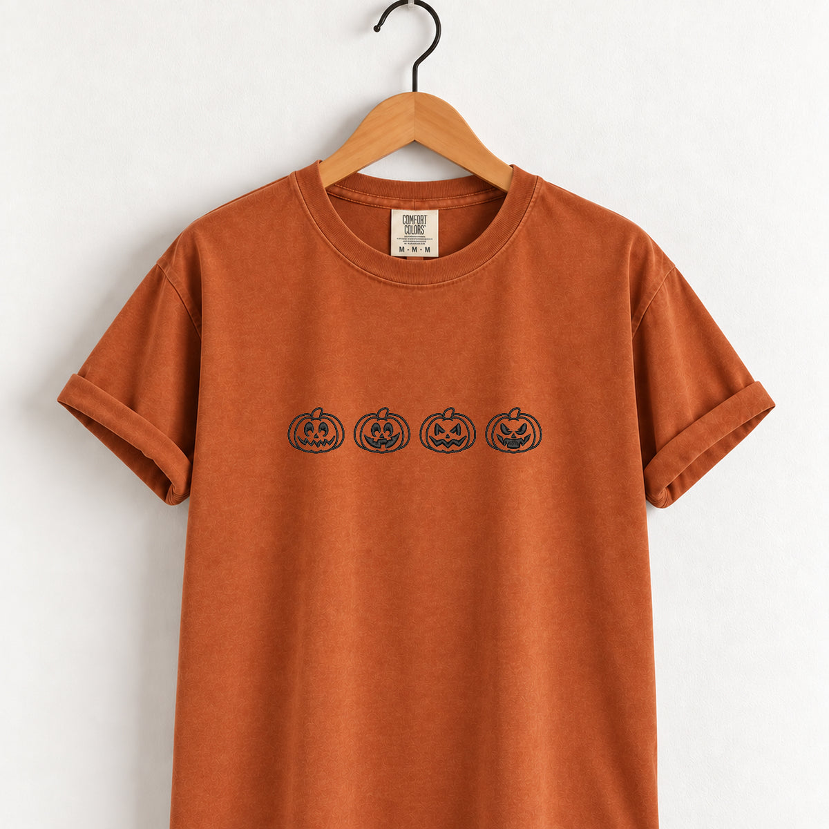 Jack-o-lantern embroidered tee in Yam on a wooden hanger, four black-thread pumpkins across the chest