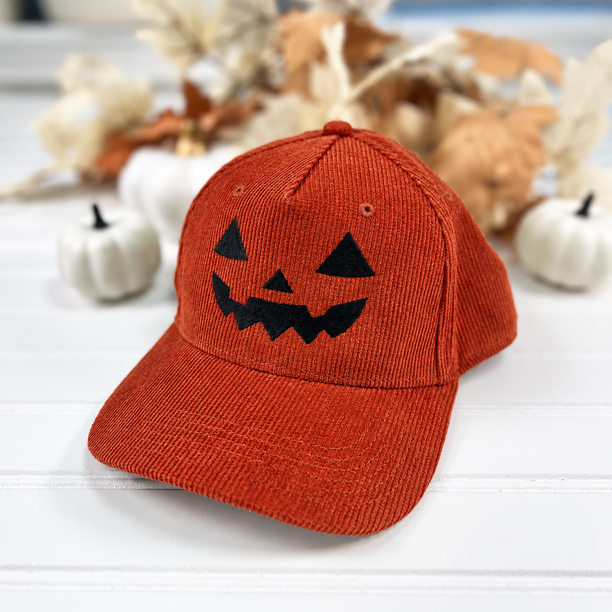 Jack-o-lantern embroidered corduroy snapback hat in Rust with a black thread pumpkin face, styled with white pumpkins and fall leaves