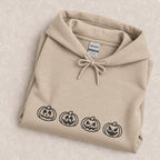 Jack-o-lantern embroidered hoodie in Sand folded on a cream floral surface, close up of the four black thread pumpkins