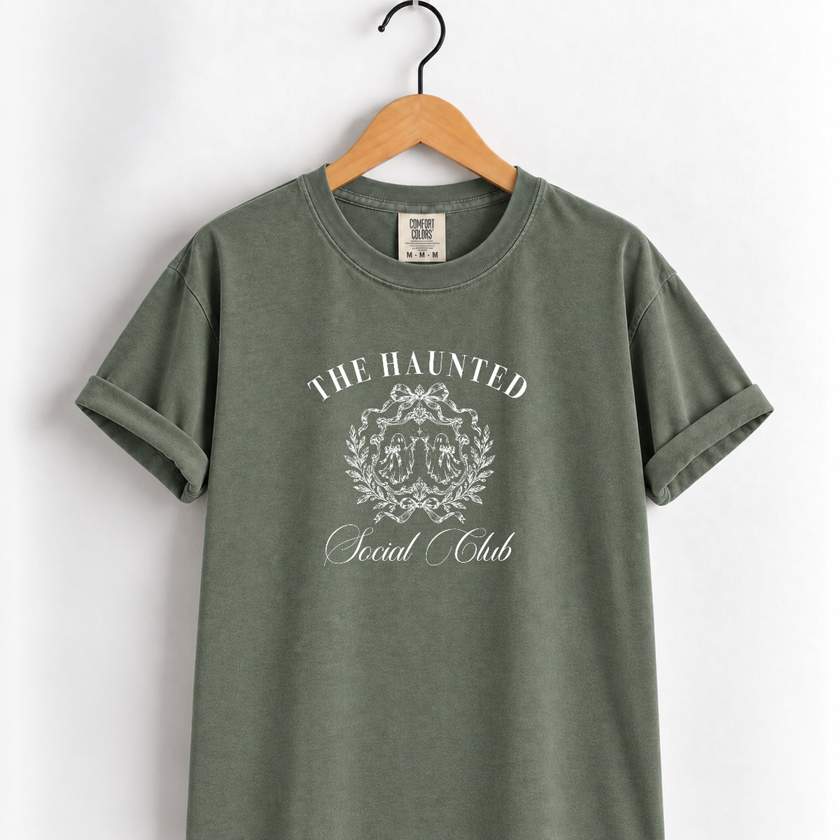 The Haunted Social Club tee in Moss on a wooden hanger, white vintage crest with ghosts and a bow