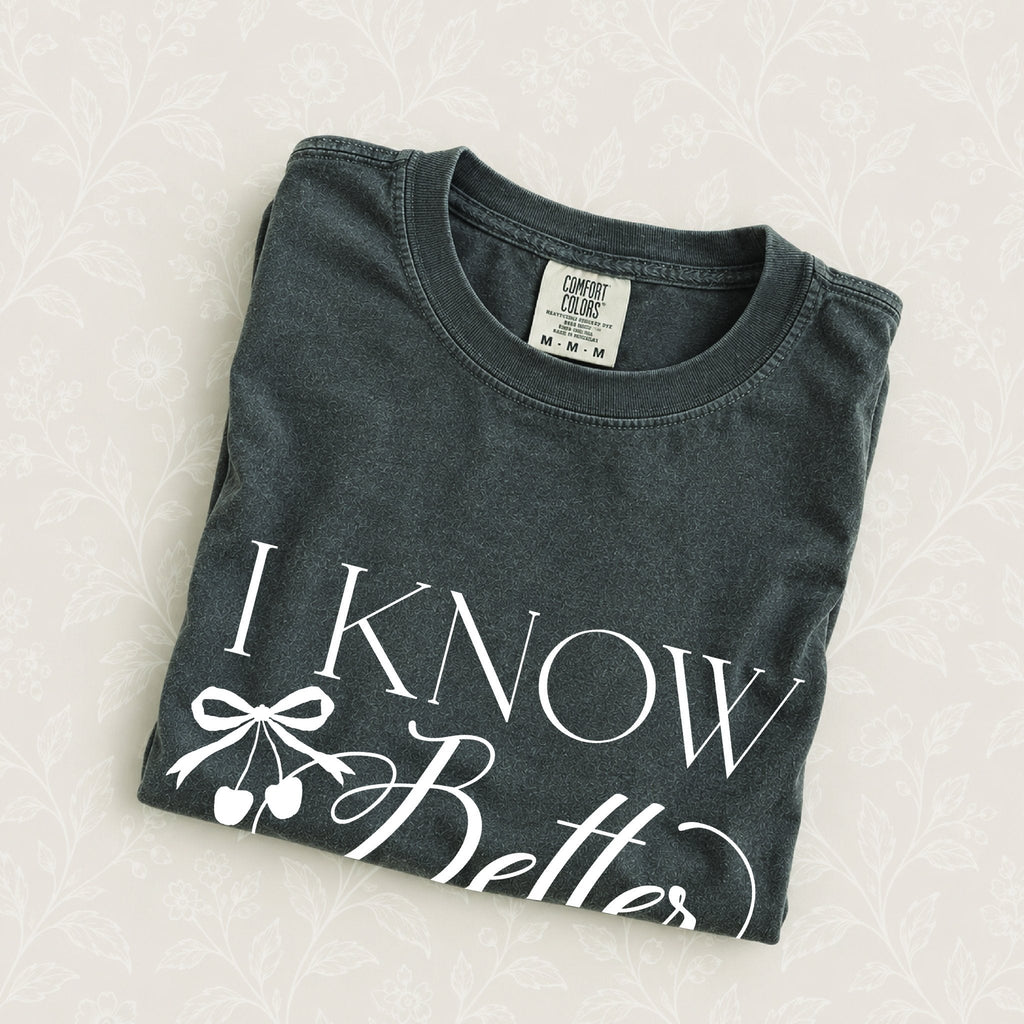I Know Better, I Simply Chose Otherwise tee in Pepper, folded on a cream floral surface, cherry bow design close up