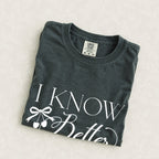I Know Better, I Simply Chose Otherwise tee in Pepper, folded on a cream floral surface, cherry bow design close up