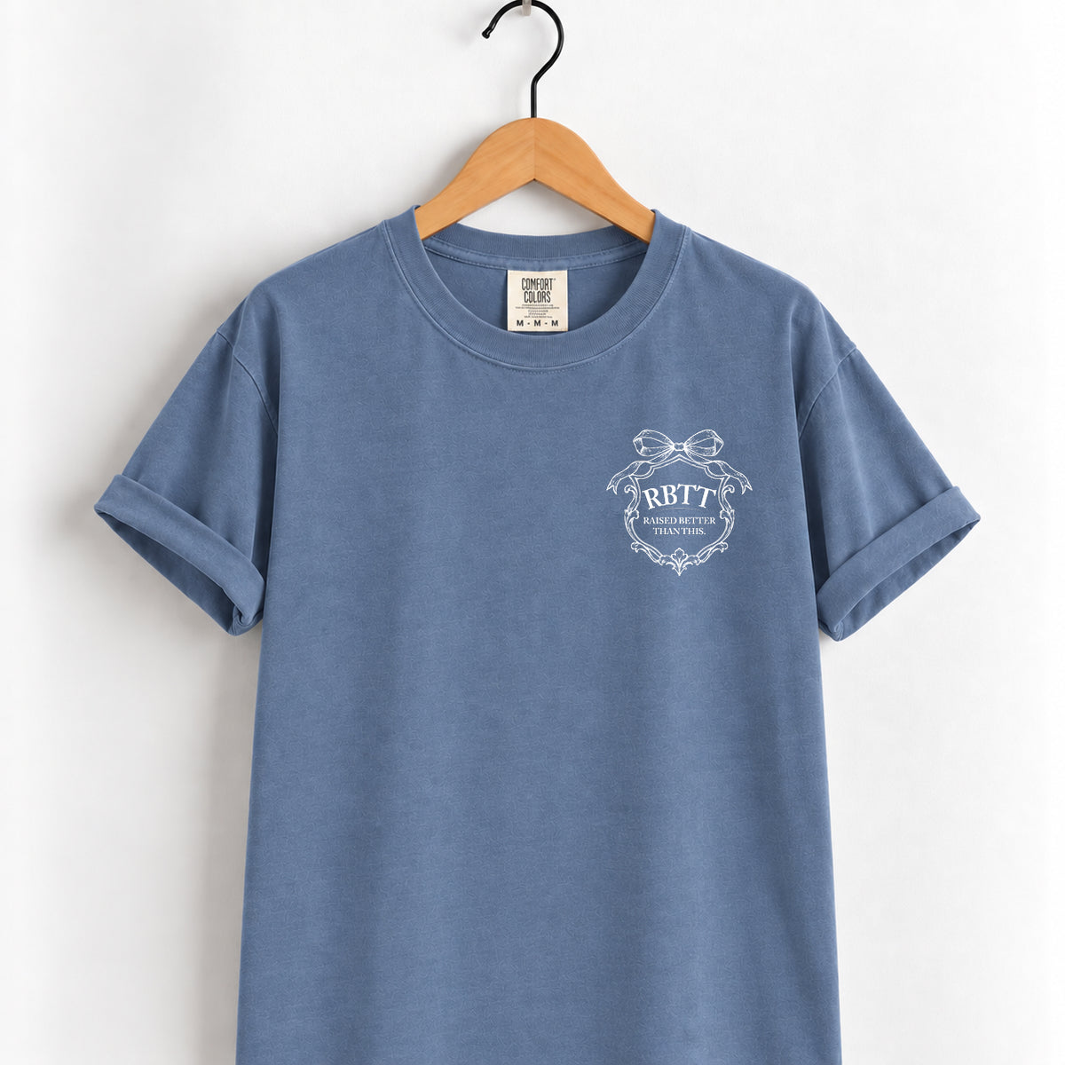 Raised Better Than This crest tee in Blue Jean on a wooden hanger, left-chest RBTT logo with bow