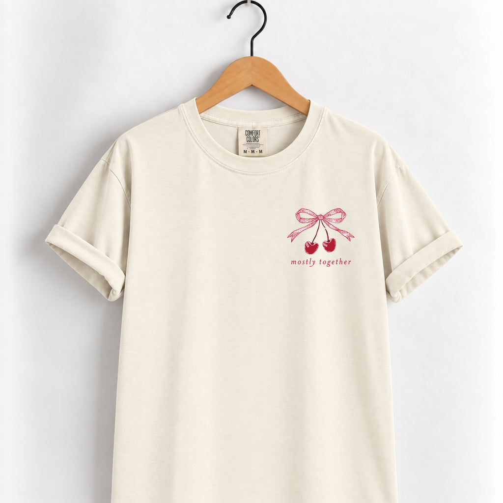 Mostly Together cherry bow tee in Ivory on a wooden hanger, left-chest print in deep red