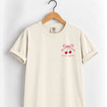 Mostly Together cherry bow tee in Ivory on a wooden hanger, left-chest print in deep red