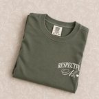 Respectfully, No tee in Moss folded on a cream floral surface, left-chest print close up