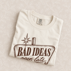 Bad Ideas, Open Late tee in Ivory folded on a cream floral surface, sign close up