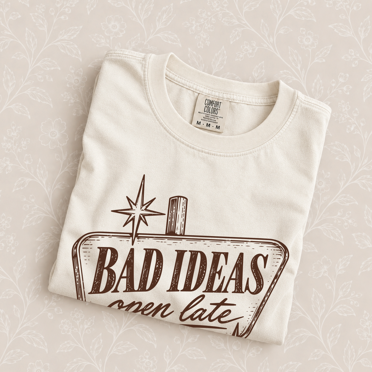 Bad Ideas, Open Late tee in Ivory folded on a cream floral surface, sign close up