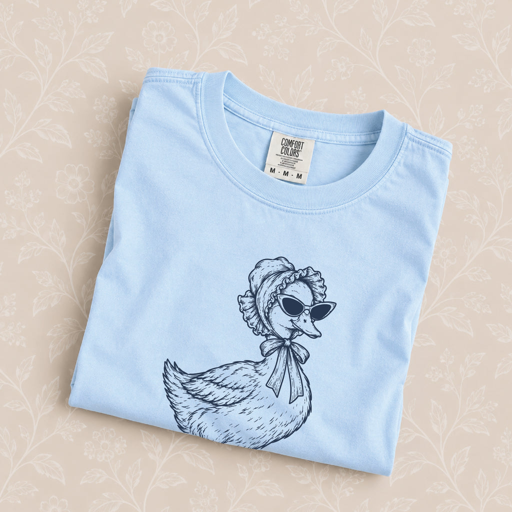 Duck Off tee in Chambray folded on a cream floral surface, close up of the duck in a bonnet and cat-eye sunglasses