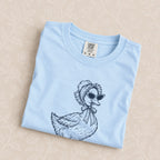 Duck Off tee in Chambray folded on a cream floral surface, close up of the duck in a bonnet and cat-eye sunglasses