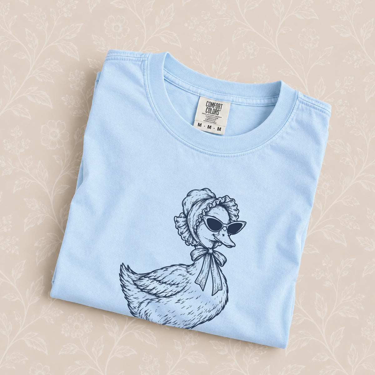 Duck Off tee in Chambray folded on a cream floral surface, close up of the duck in a bonnet and cat-eye sunglasses